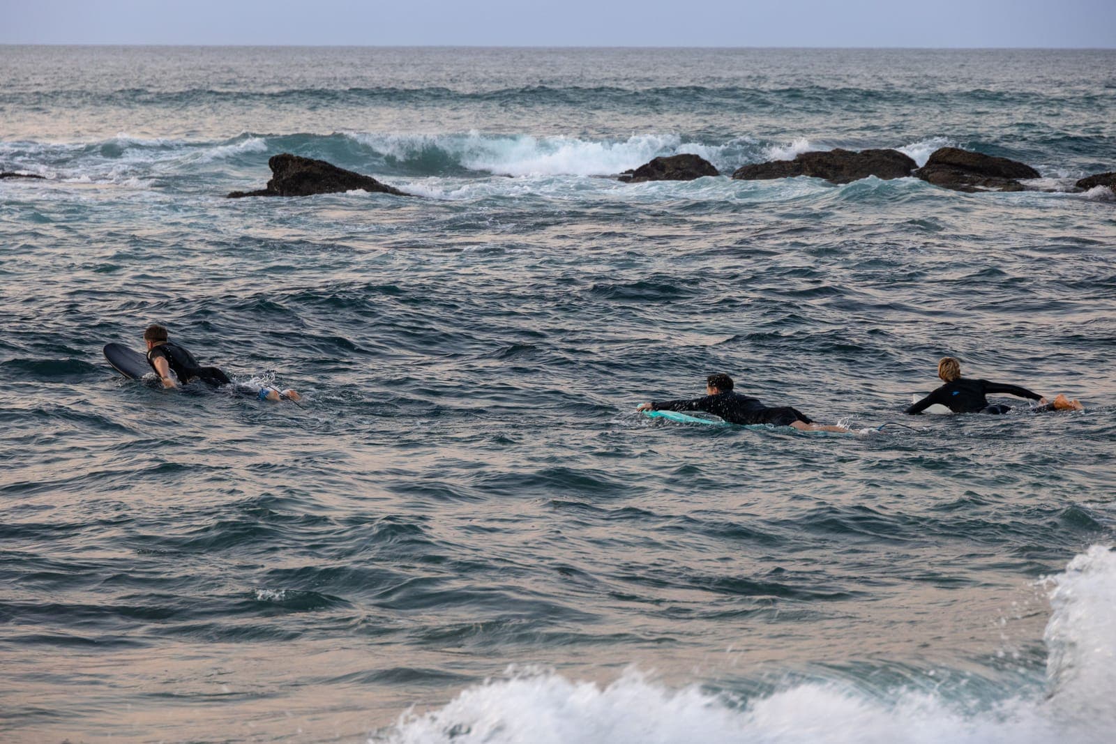 Three surfers in wetsuits paddling out past rocks on a choppy sea