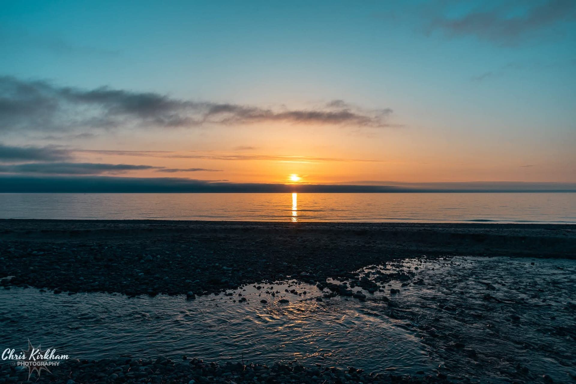 The sun setting over a calm sea, seen from a pebble shore with a stream running into it
